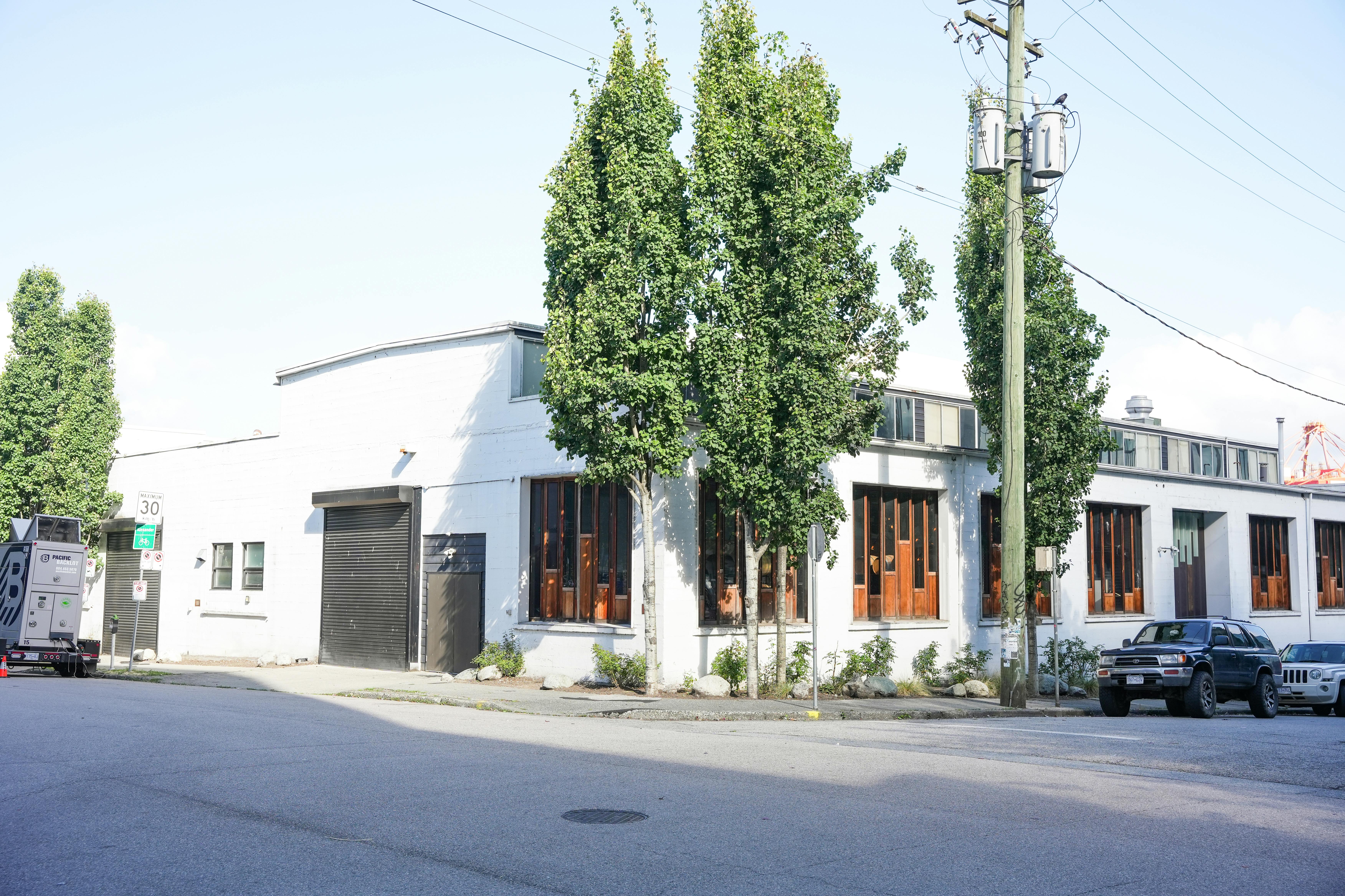 Single-story white industrial building with tall narrow wood-framed windows and a black garage door. Several trees line the sidewalk, and parked vehicles are visible along the street.