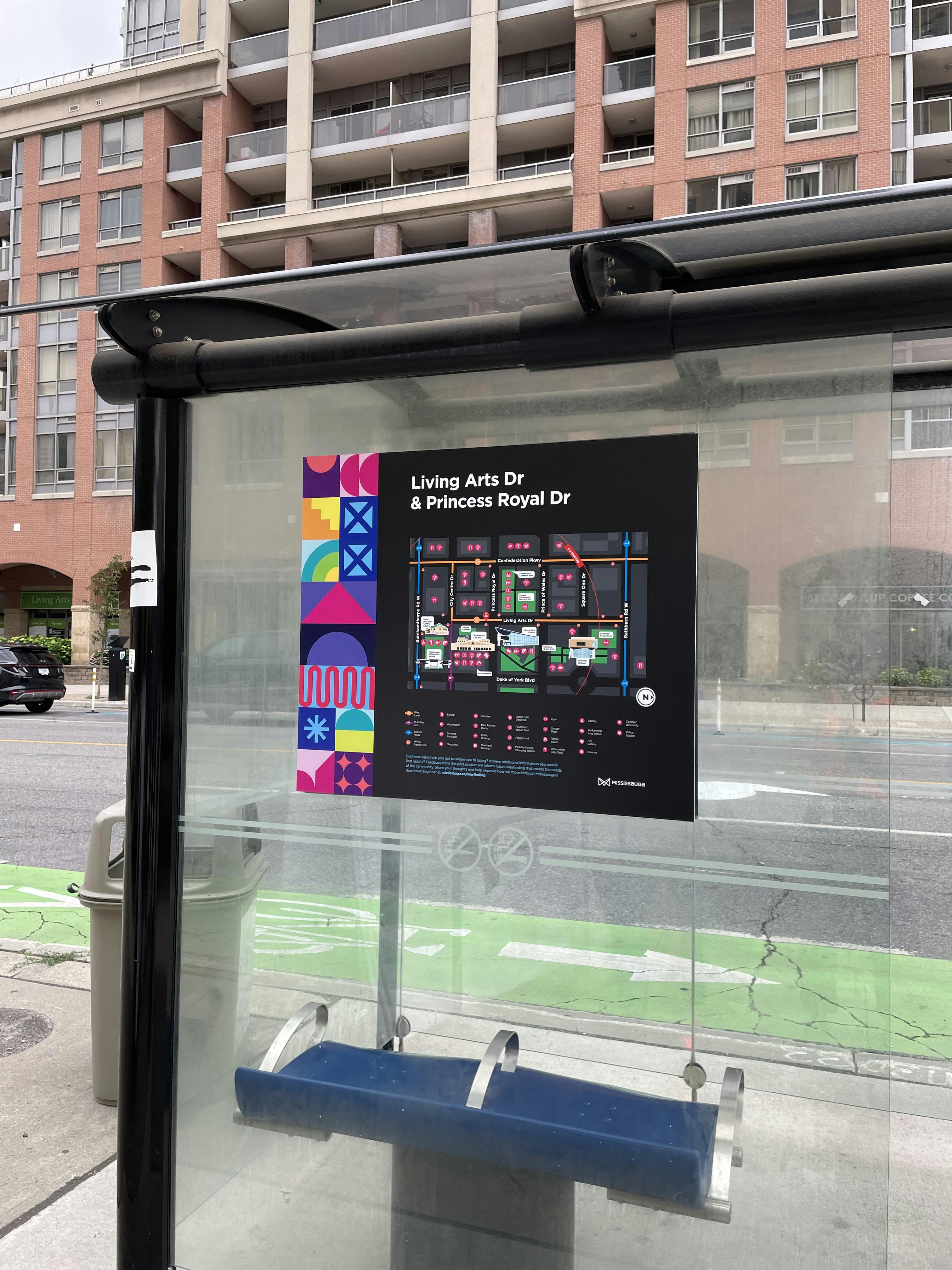 A transit shelter with a colorful map showing Living Arts Dr and Princess Royal Dr; apartment building and street visible in the background.
