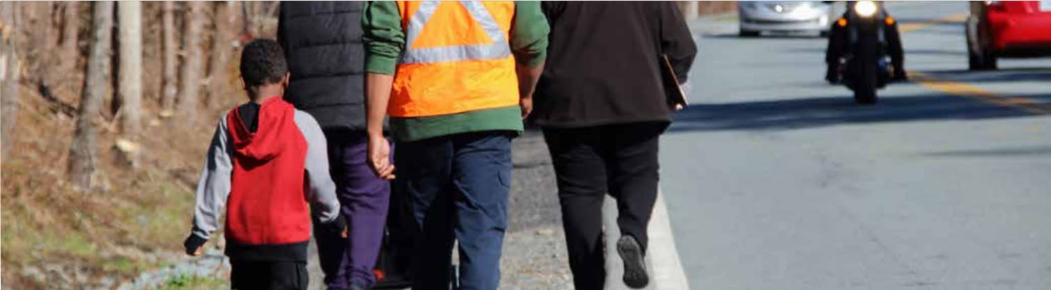 One child and three adults are walking beside a road that does not have a sidewalk. 