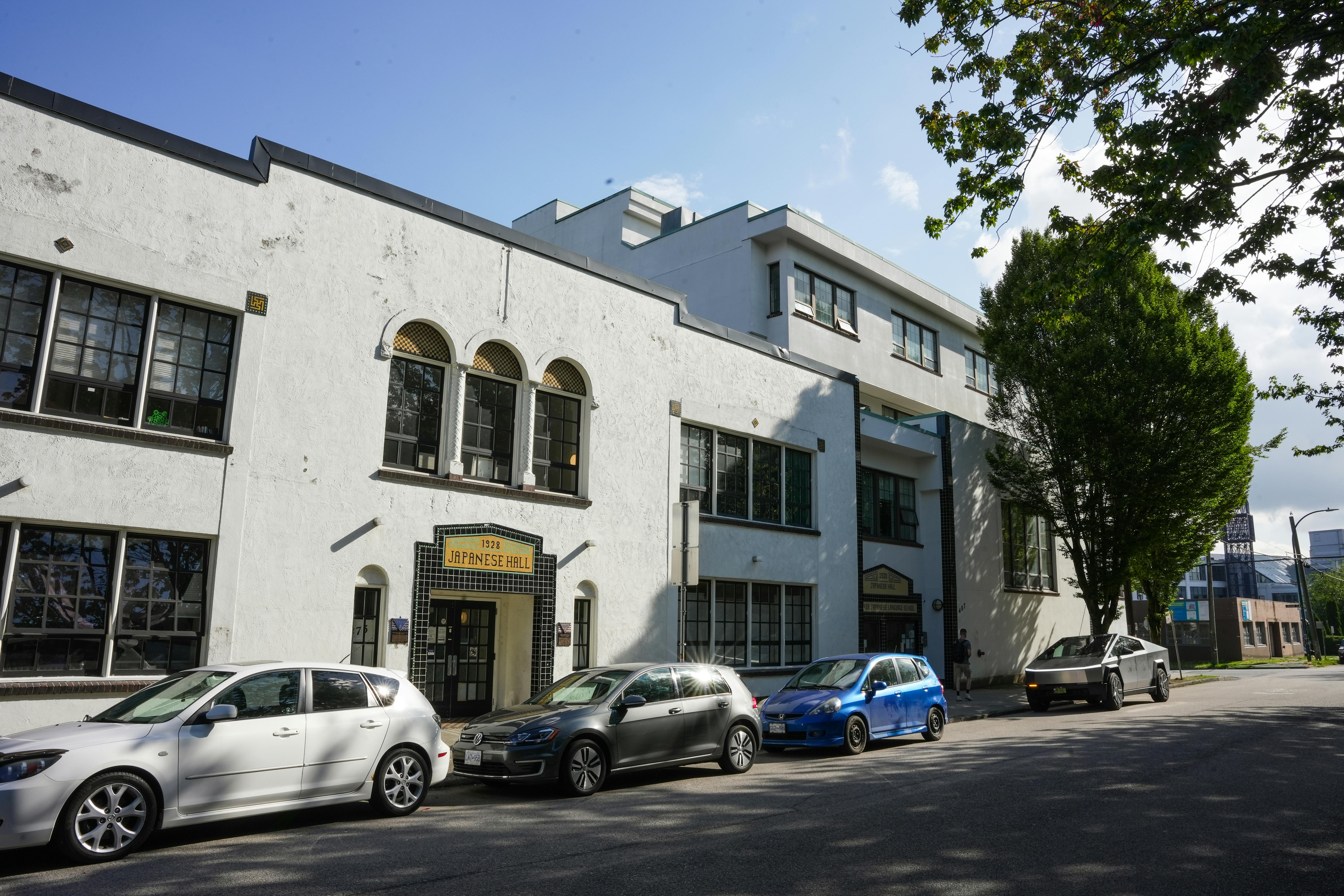 Historic Japanese Language School and Hall building with white stucco exterior. Several cars are parked along the street, and a large tree partially shades the building.