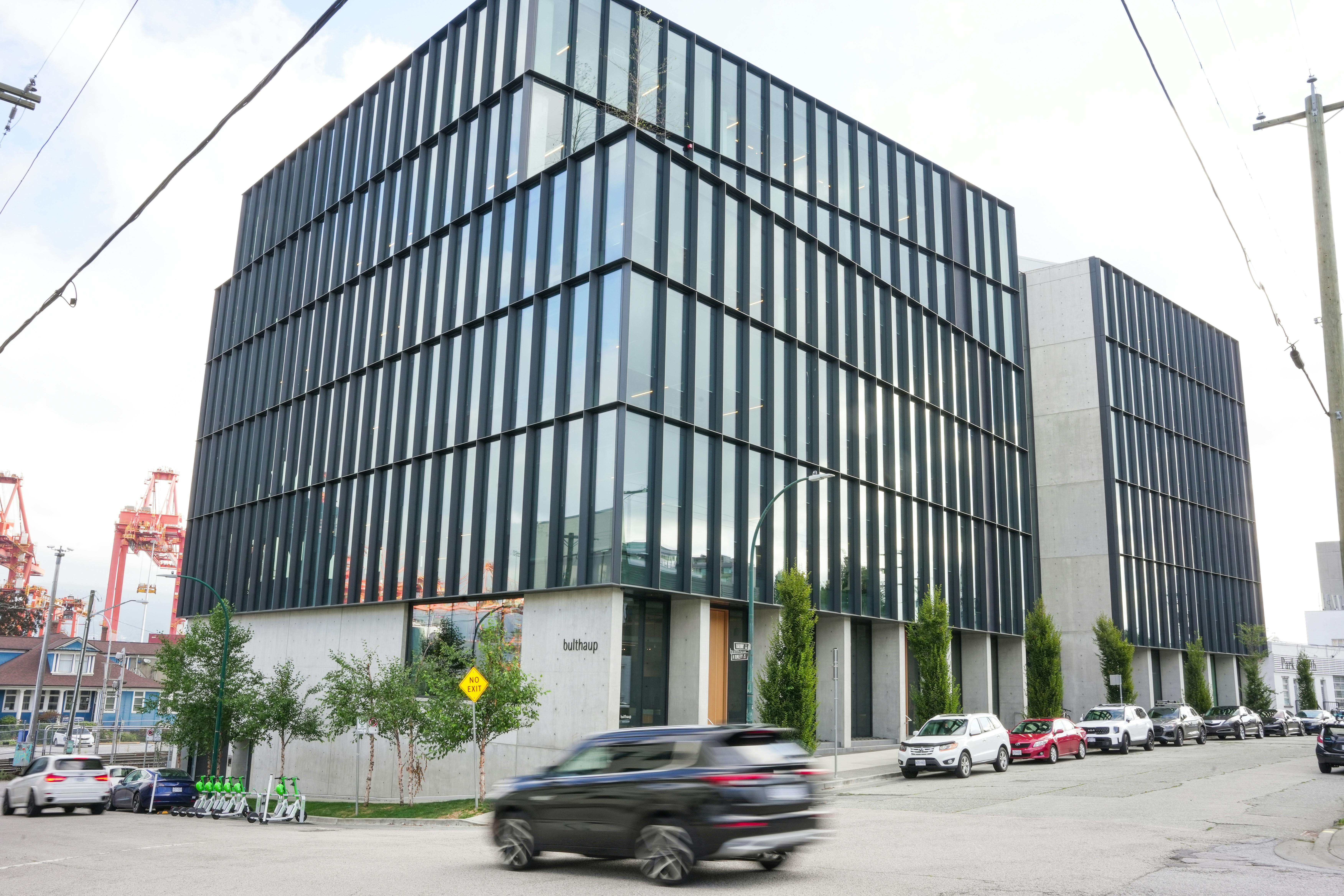 Modern office building with dark vertical glass panels and a concrete base. The structure has a sleek, contemporary design with small trees planted along the sidewalk and parked cars lining the street. Industrial cranes are visible in the background.