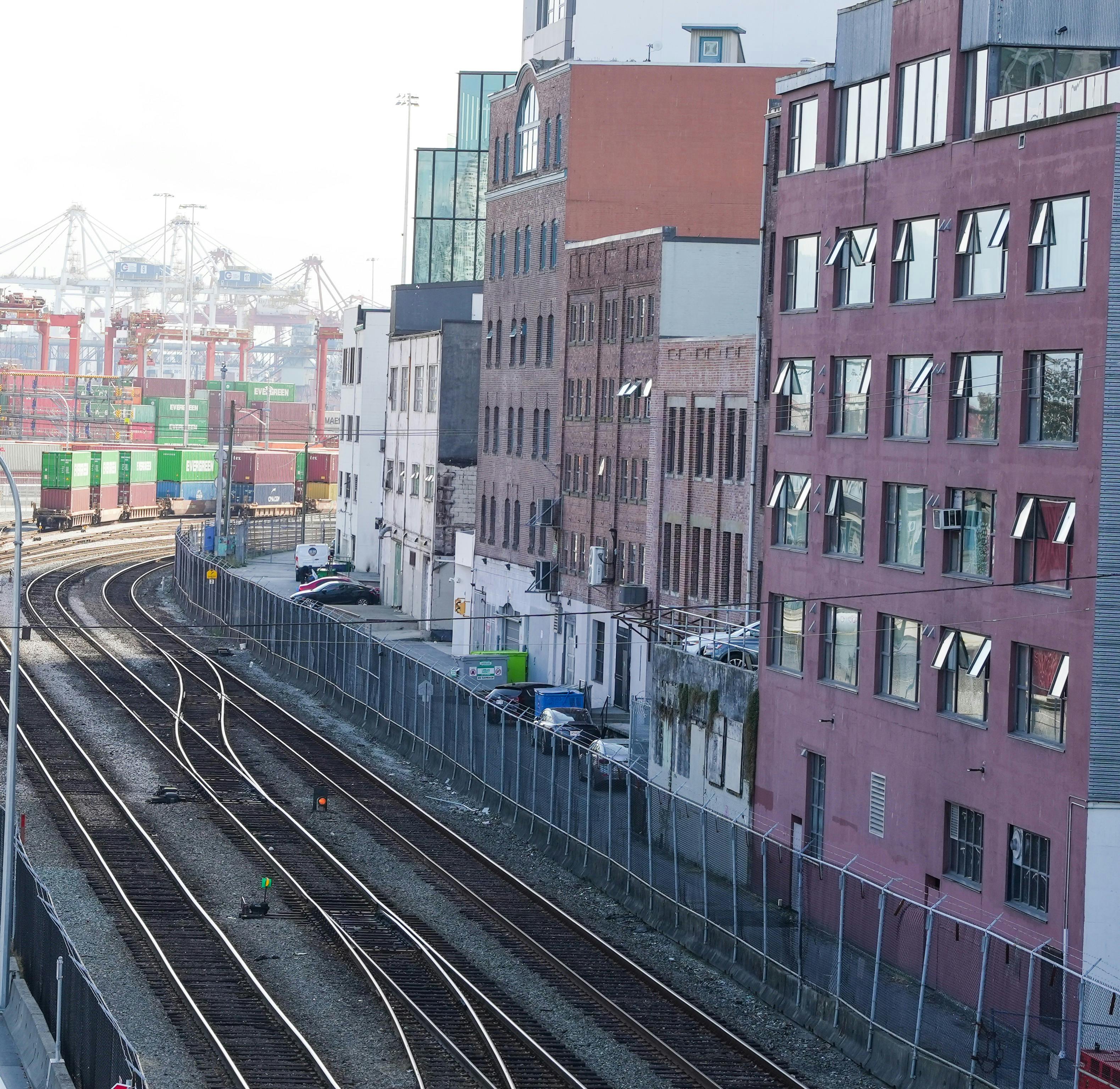 Row of multi-story brick and painted buildings with windows along railway tracks. A narrow road with parked cars runs beside the buildings, separated from the tracks by a chain-link fence. Shipping containers and cranes are in the background.