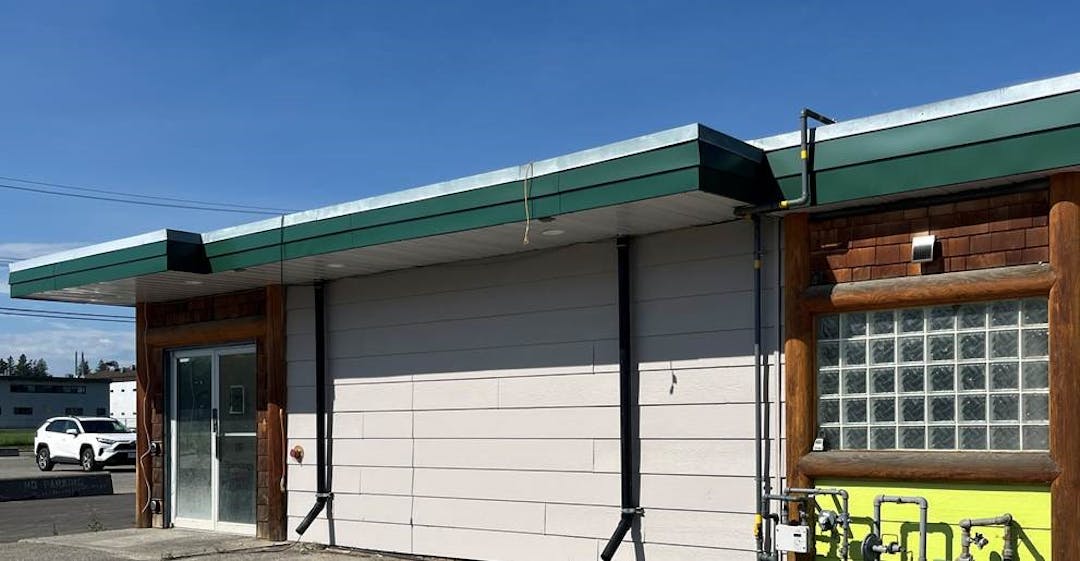 Single-story building with green trim, glass door and window, utility meters, and a white car under blue sky.