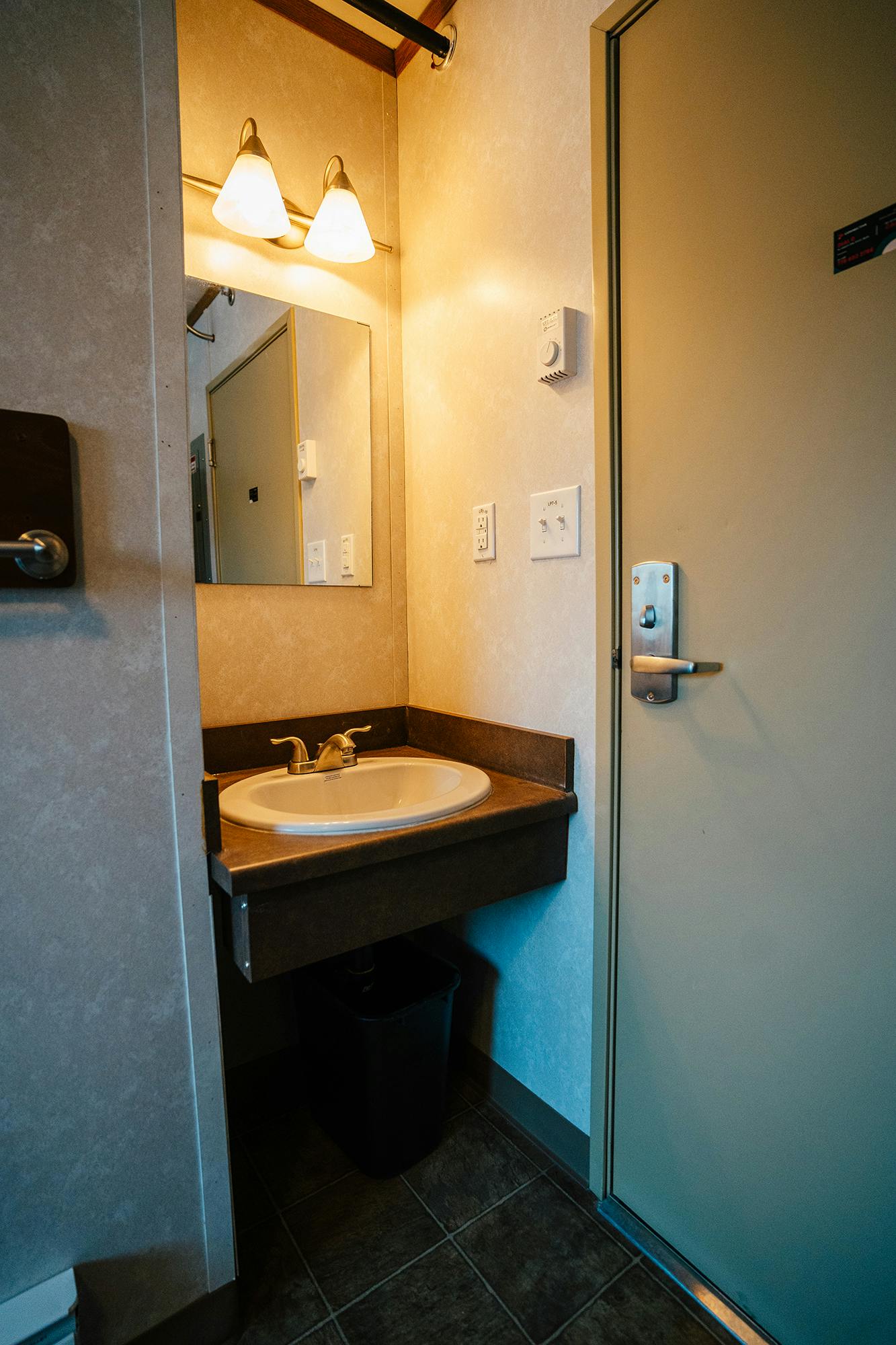 A compact bathroom with a sink, mirror, and a door, illuminated by a wall-mounted light fixture.