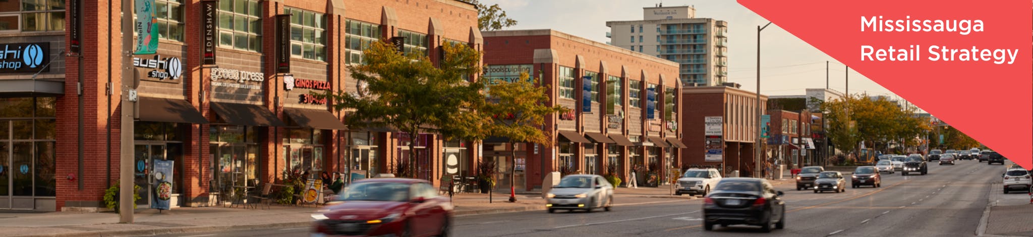 Picture of a sunny streetscape with cars driving by in the foreground with shops in the middle ground