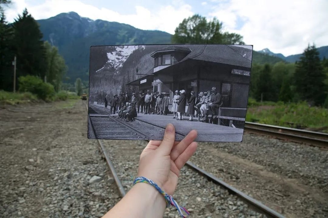 A hand holding a black and white photo of a railway station in front of the same location in present day.