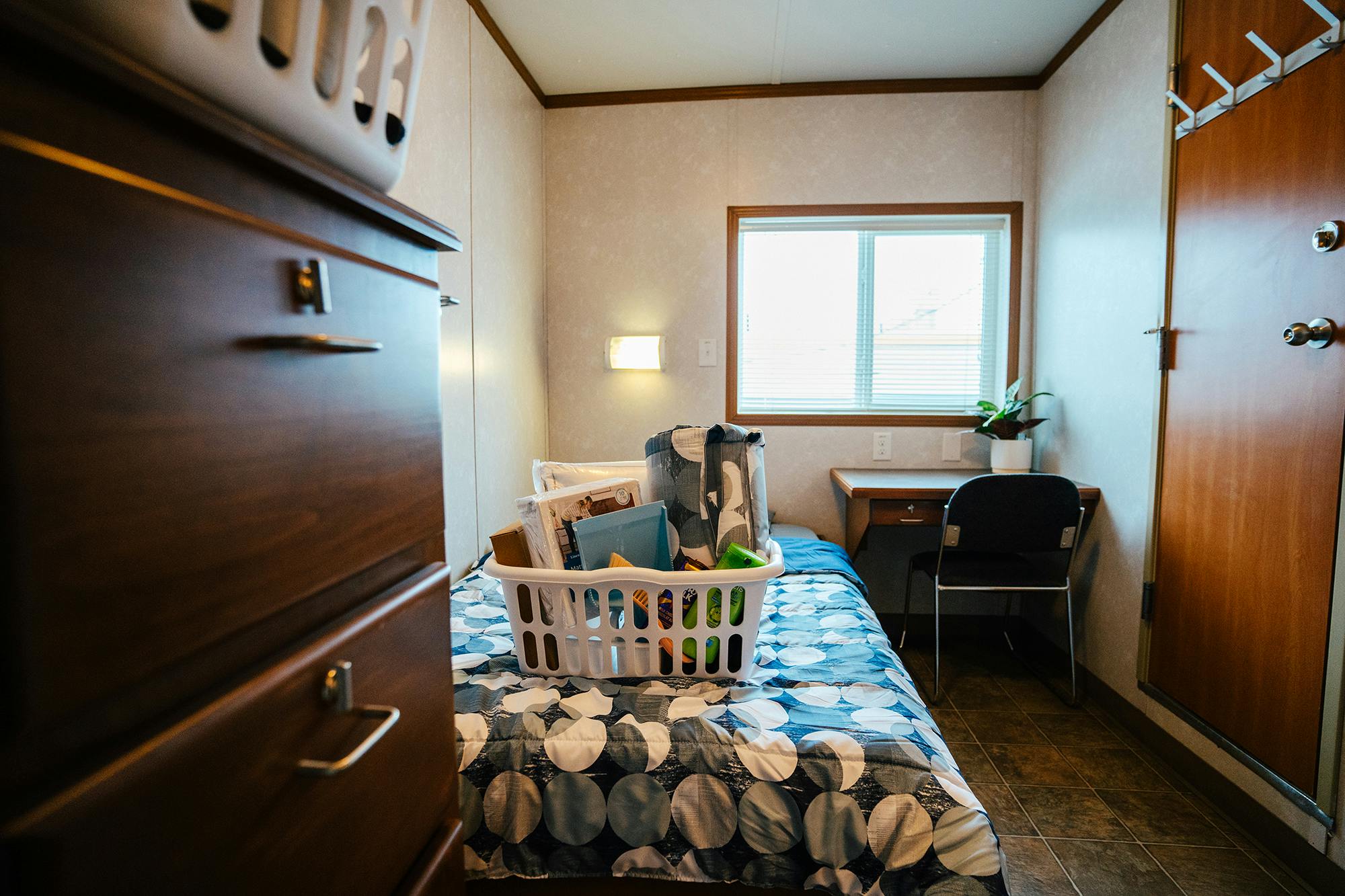 Another view of the small bedroom, featuring a single bed, a desk under the window, a wooden dresser, and a laundry basket filled with essential items.