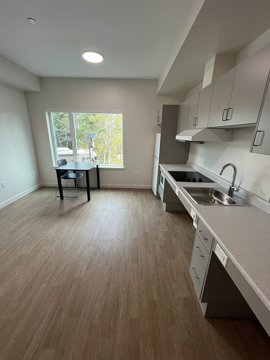 A modern kitchen and dining area with white cabinets, light wood flooring, and a small black table with two chairs near a large window overlooking greenery.
