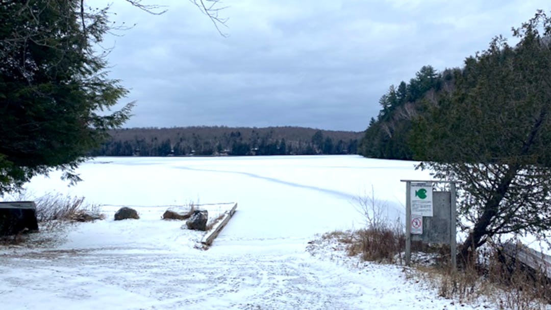 Image of a frozen lake, trees surrounding it, with a wooden retaining wall on the left of a boat launch under grey sky