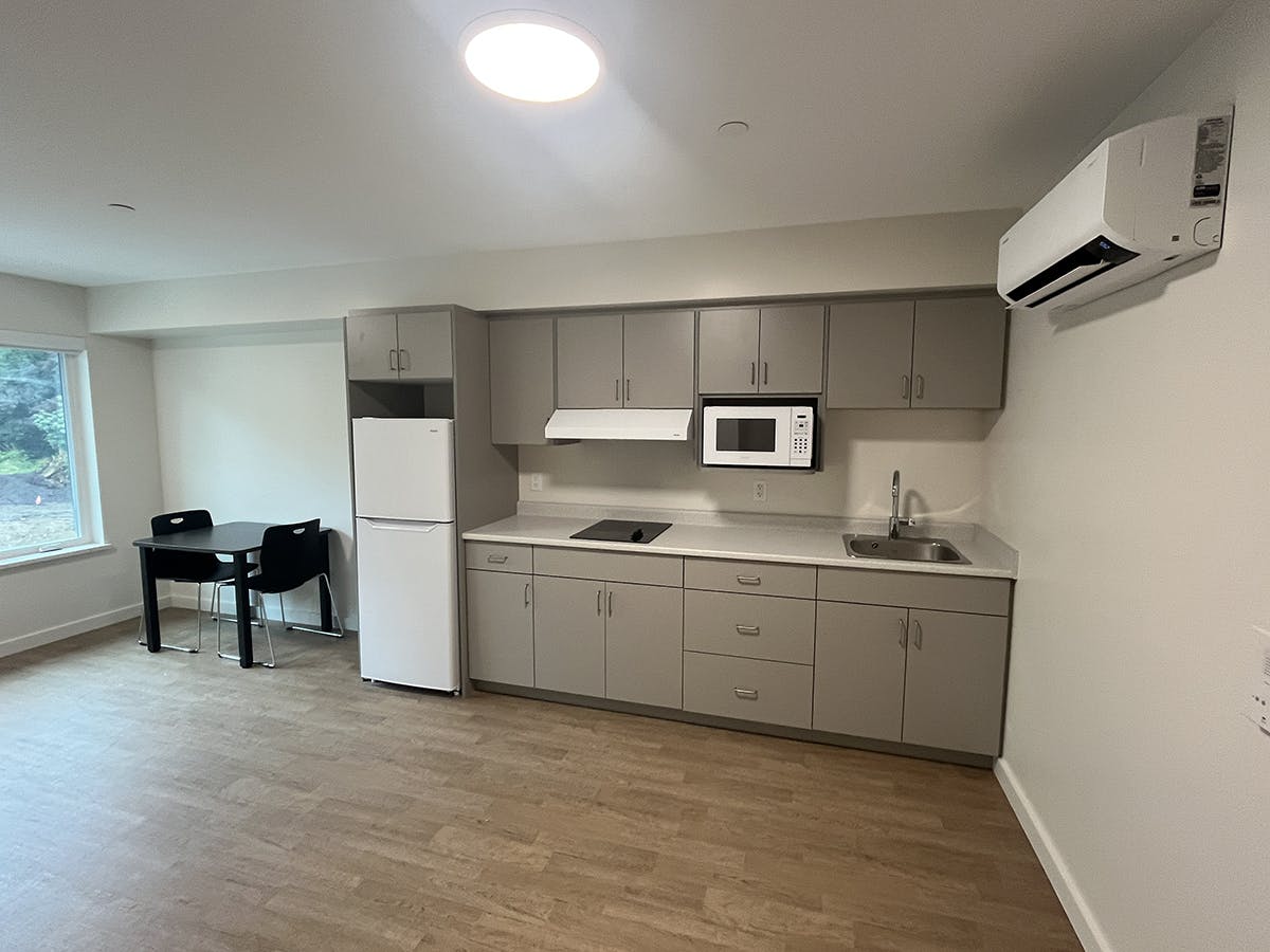 A modern kitchen with grey cabinets, white appliances, and light wood flooring, featuring a small dining area with a black table and chairs near a window and an air conditioning unit on the wall.