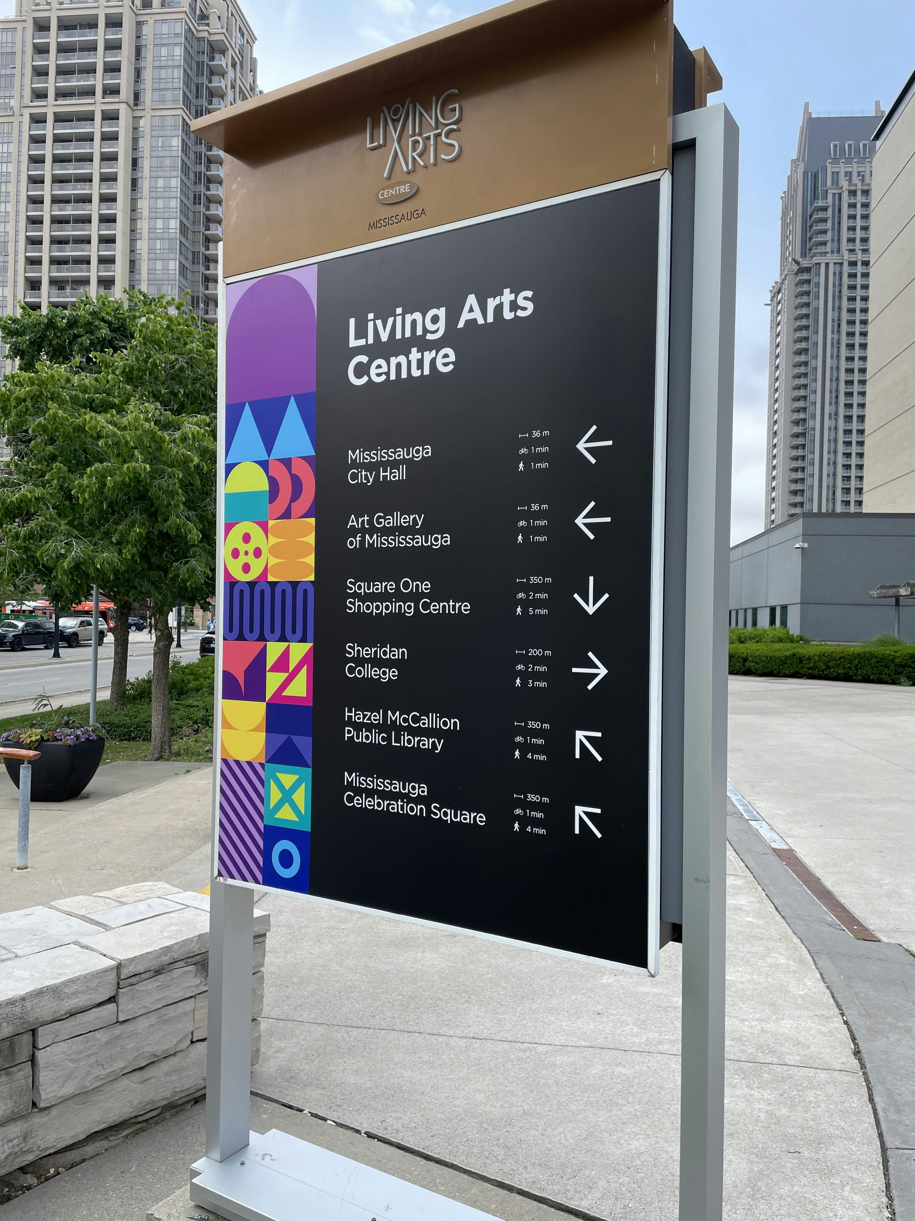 Directional sign for Living Arts Centre in Mississauga, listing nearby locations with arrows and distances, against an urban backdrop with tall buildings and greenery.