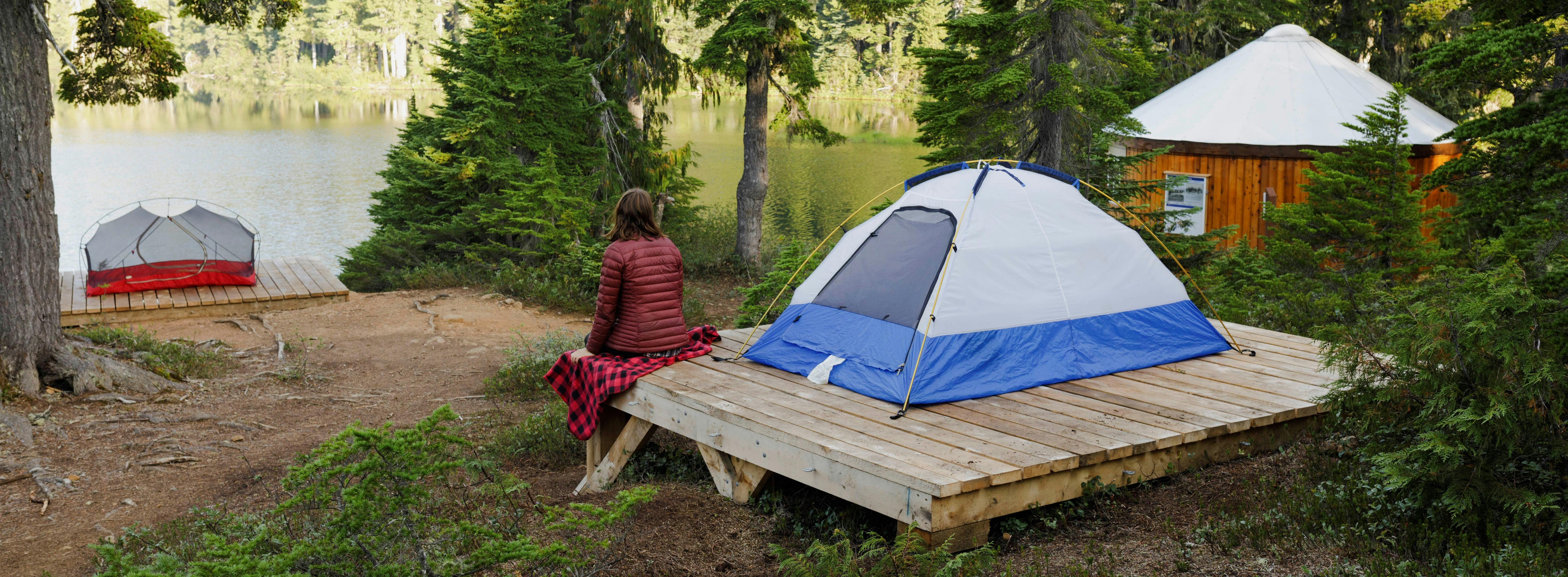 A person sits on the corner of a tent platform looking at a lake in the distance. (Croteau Lake, Strathcona Park)