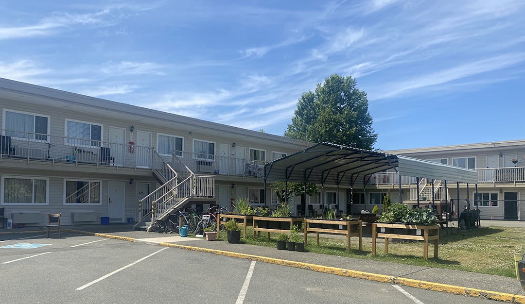 Two-story motel with exterior stairs and balconies, courtyard seating, potted plants, and bikes under a sunny sky.