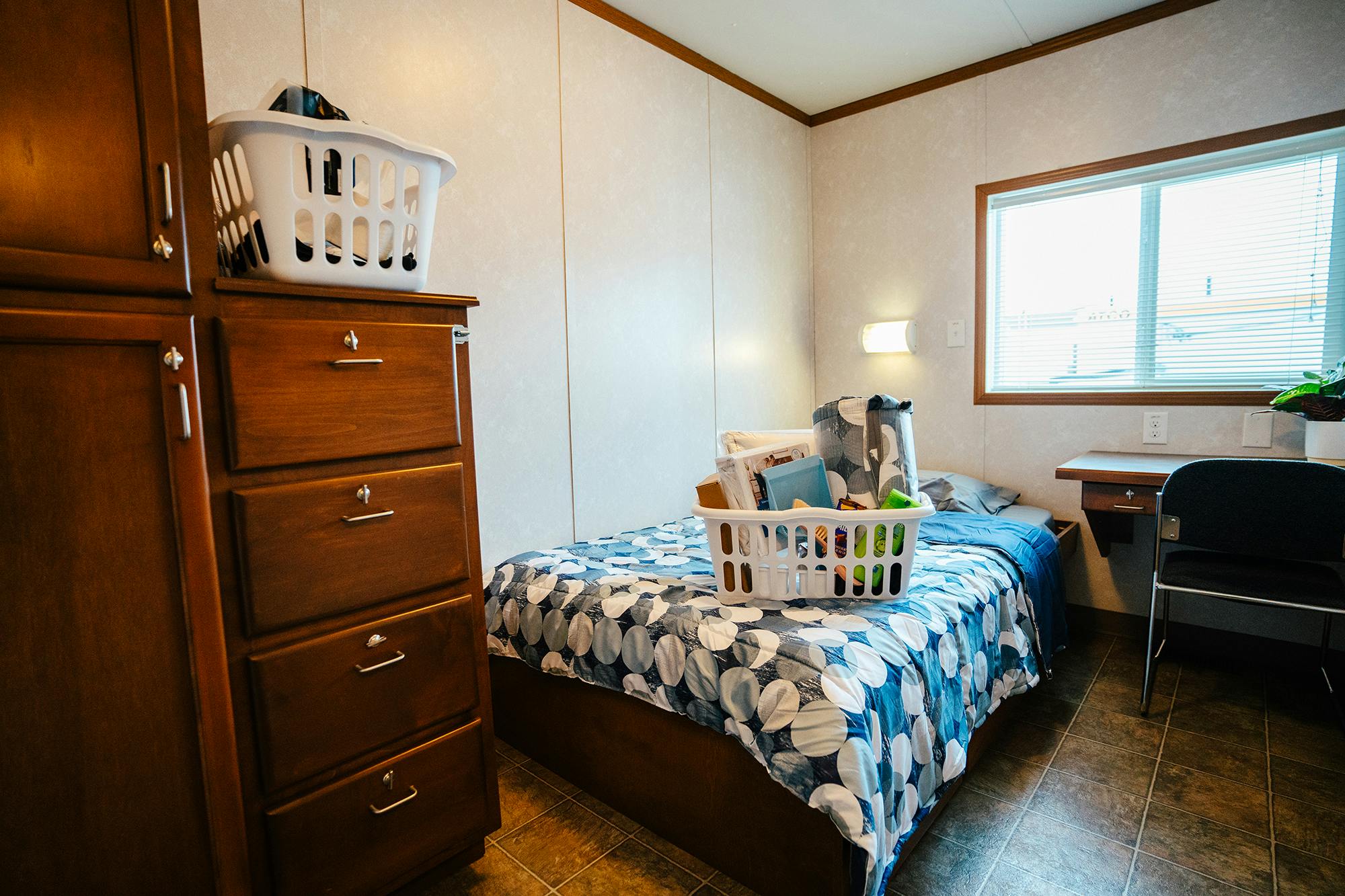 A small bedroom with a single bed, a wooden dresser, a window, and a desk, with a laundry basket filled with personal items on the bed.