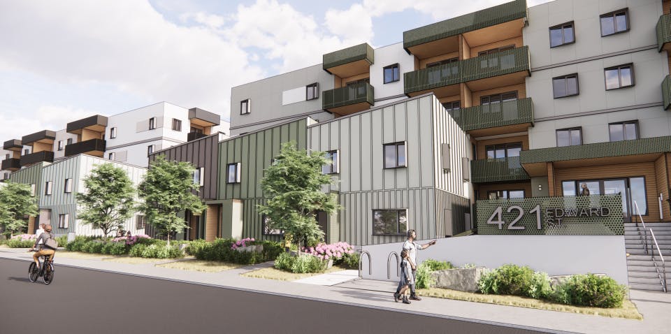 Street-level view of a modern residential building with green and brown panel siding, large windows, and landscaped greenery including trees and shrubs. A few people are walking and cycling along the sidewalk in front of the building.