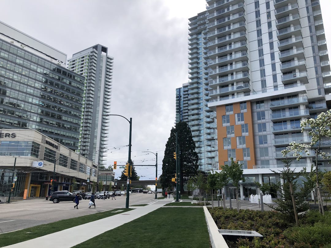 A photo from SW Marine Drive looking west. It shows green landscaping and a tree in the foreground and high-rises in the midground. People are crossing the street.