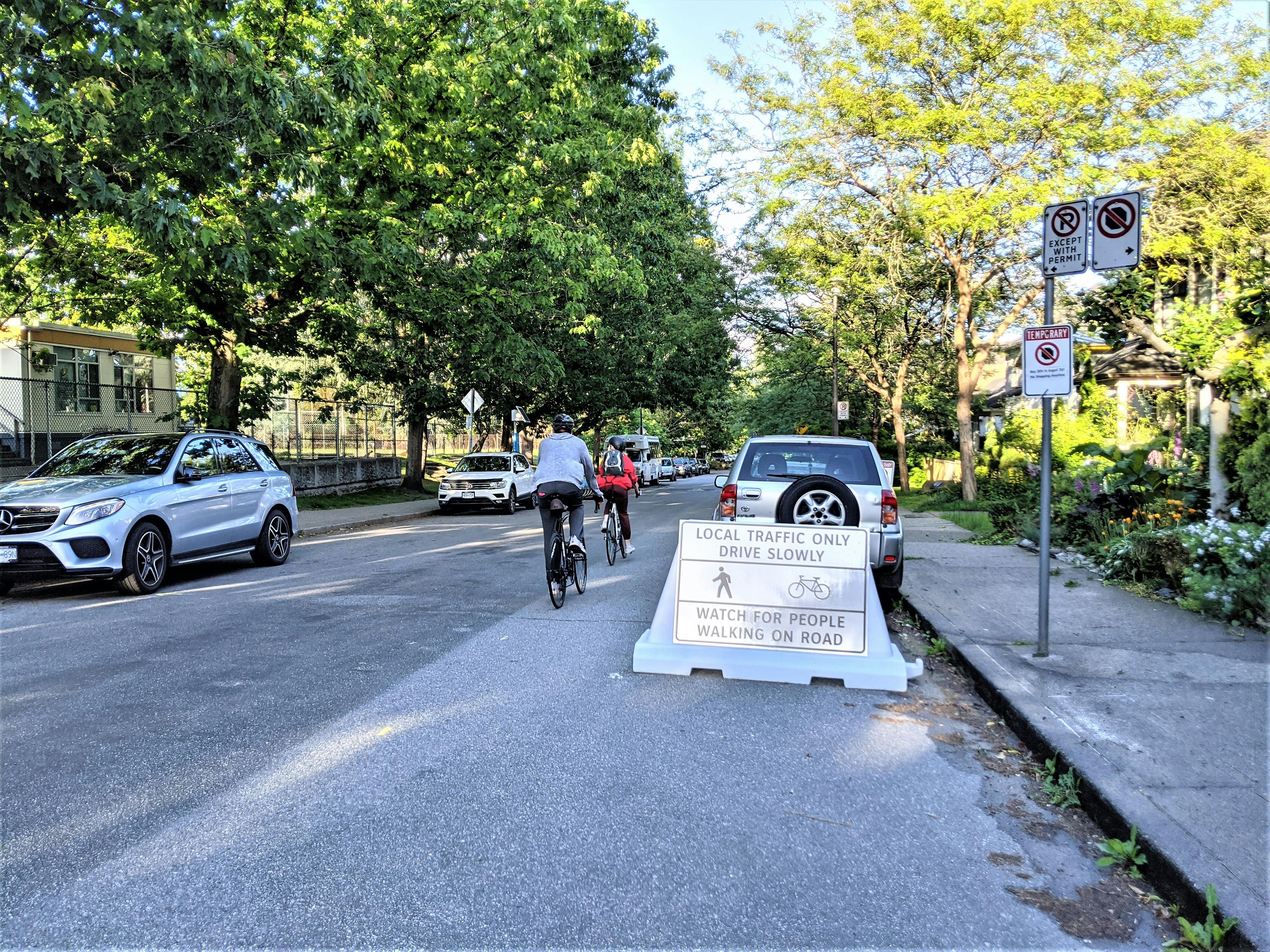 Two cyclist crossing a Slow Streets signage placed on Keefer Street. The signage reads - Local traffic only, drive slowly, watch for people walking on road along with walk and cycle icons.