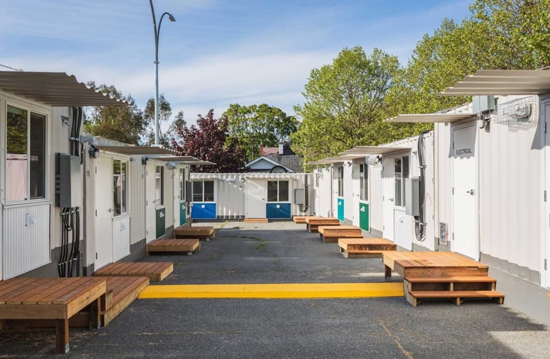 Small modular housing units line a central courtyard with wooden steps, trees, and a clear blue sky overhead.