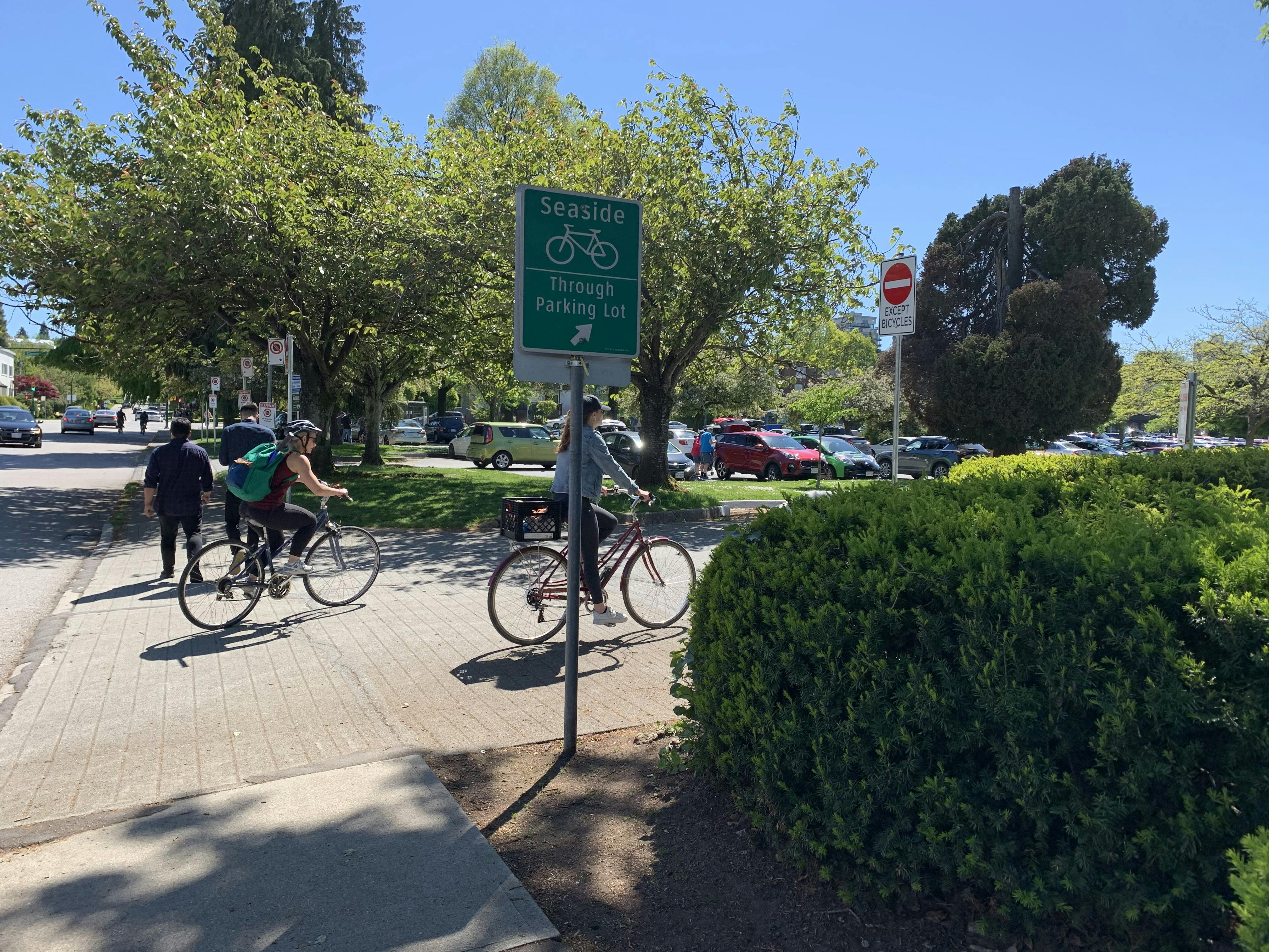 Two people cycling west into the parking lot of Kitsilano Beach Park behind a Seaside Greenway sign