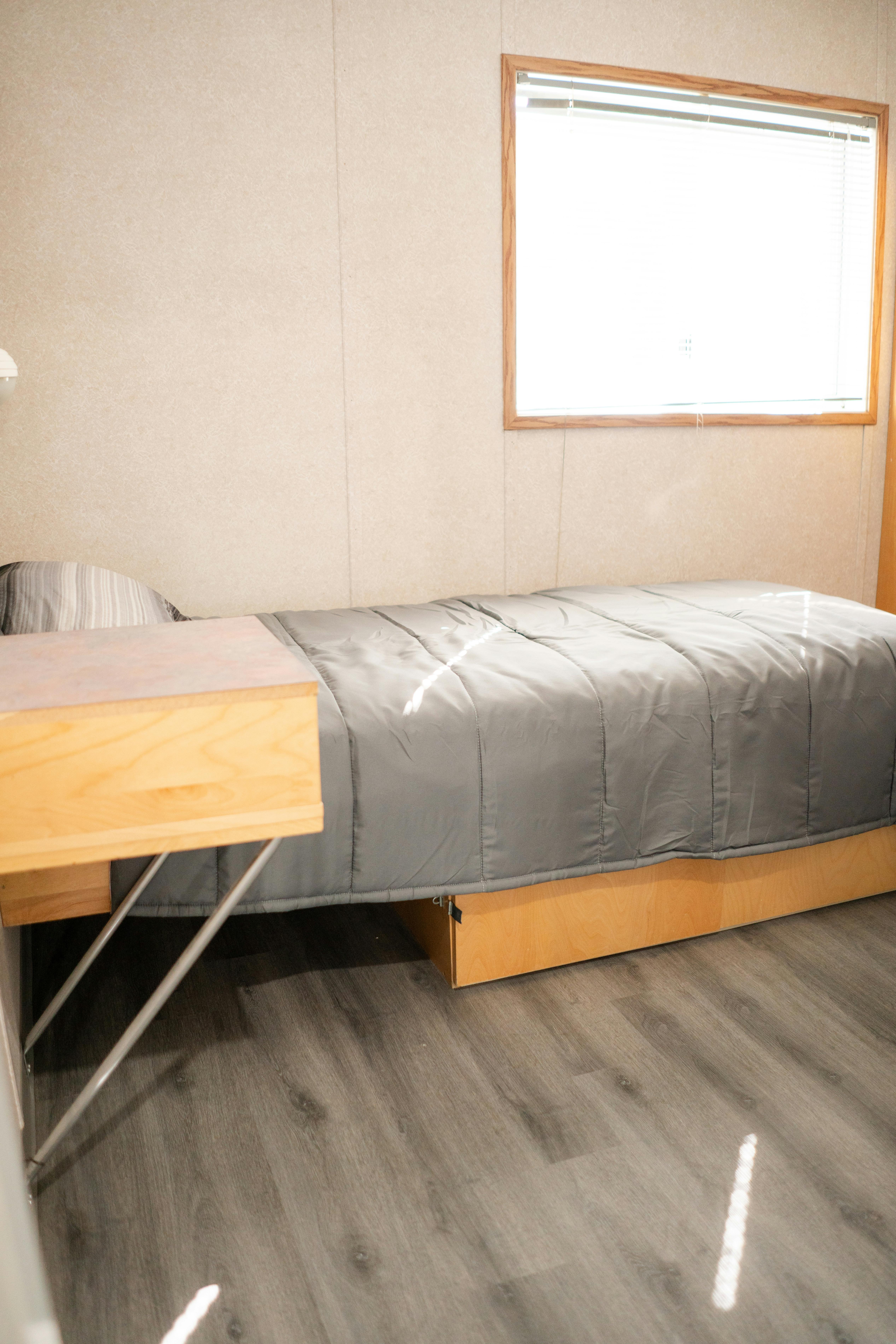 Single bed with wooden headboard, grey comforter, light brown walls, grey floor, window with blinds.