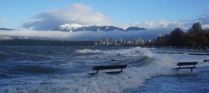 Waves crash onto a flooded beach, covering benches with water. Vancouver's downtown skyline and snowy mountains are in the background.