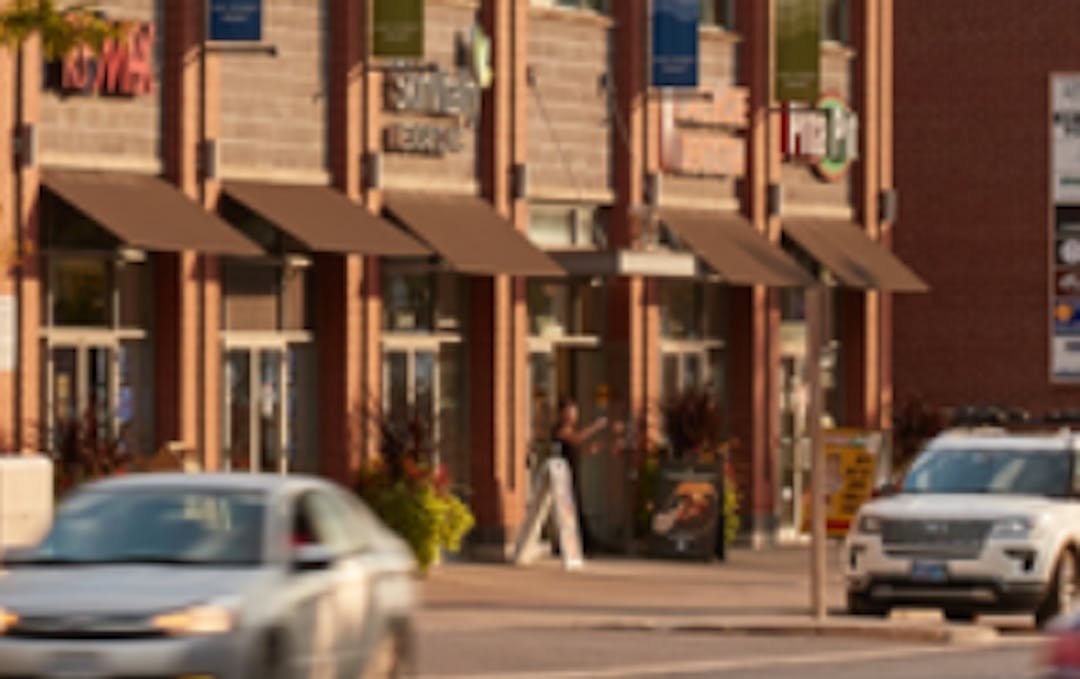 Picture of a streetscape on a sunny da with cars passing in the foreground and shops in the middle ground