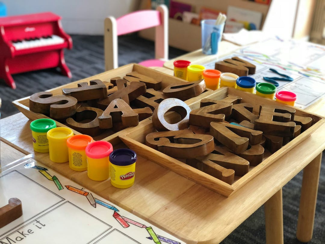 A wooden tray filled with colorful playdough and wooden letter blocks on a table in a preschool classroom.