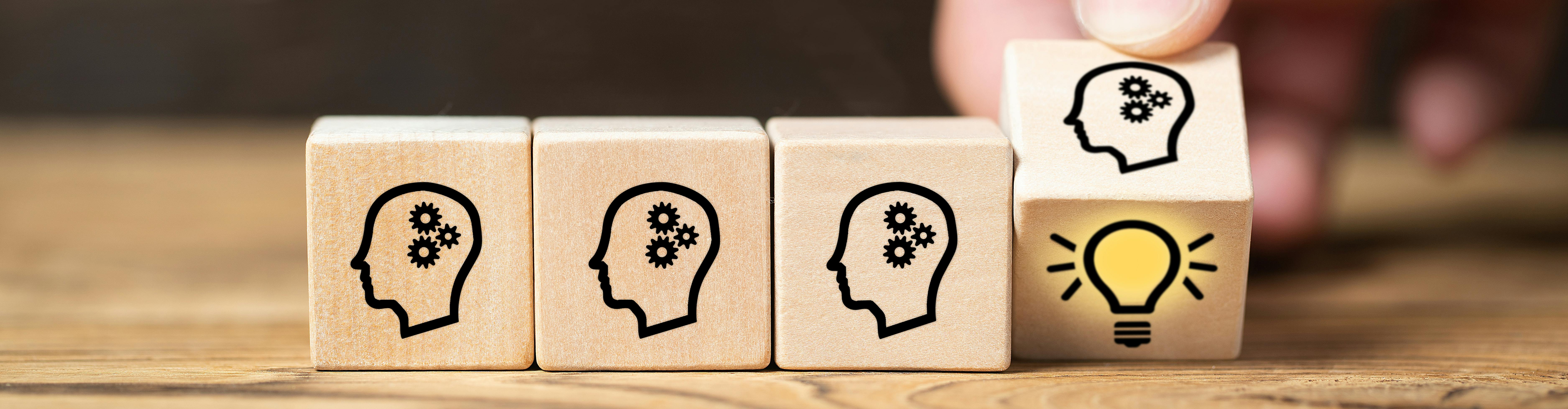 Image of wooden blocks with thinking heads printed on them. One is being turned over with the image of a yellow light bulb under it.