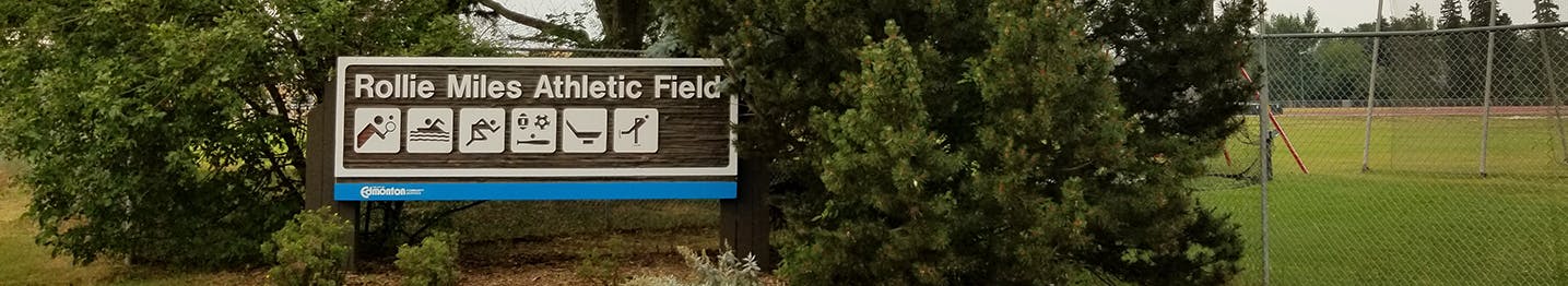 A colour image of Rollie Miles Athletic Park, with the park sign in the foreground and trees and fields in the background
