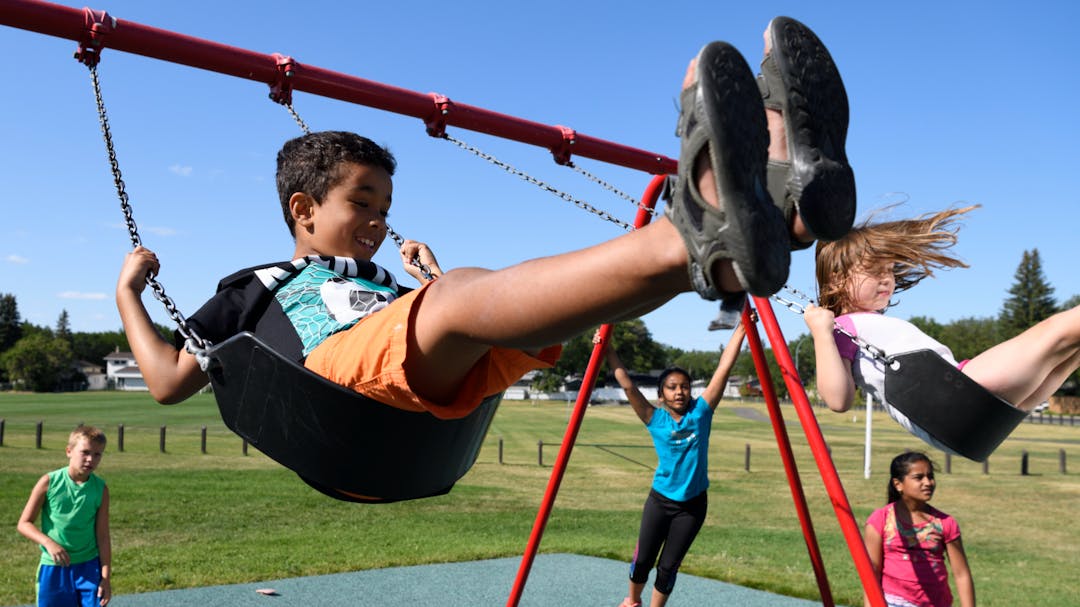 Two children swinging on swings at a playground. A few other children standing by, watching.