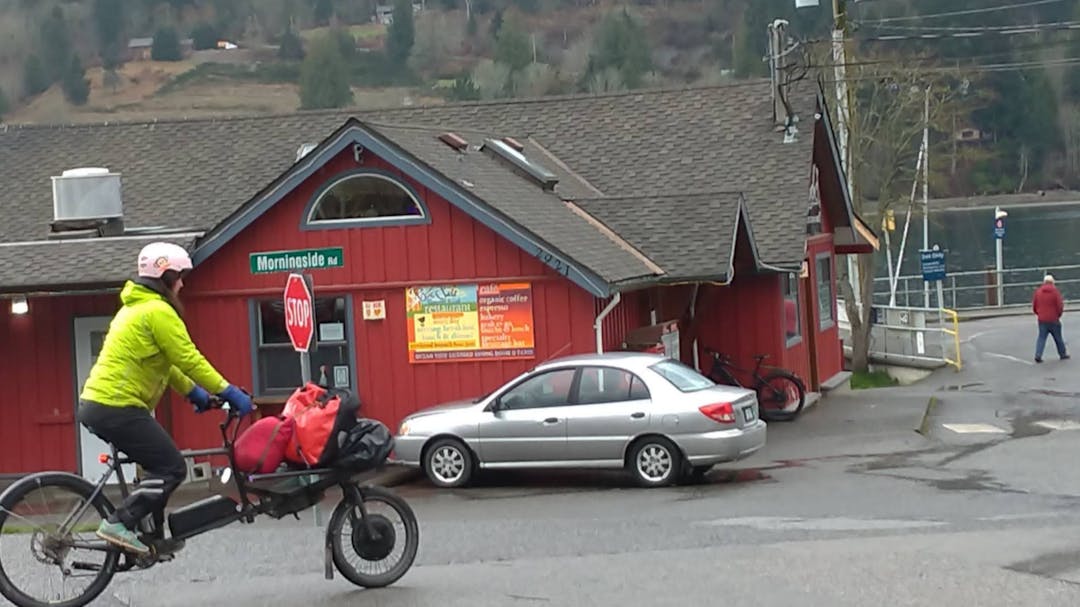 cargo bicycle riding past red building with a parked car and pedestrian