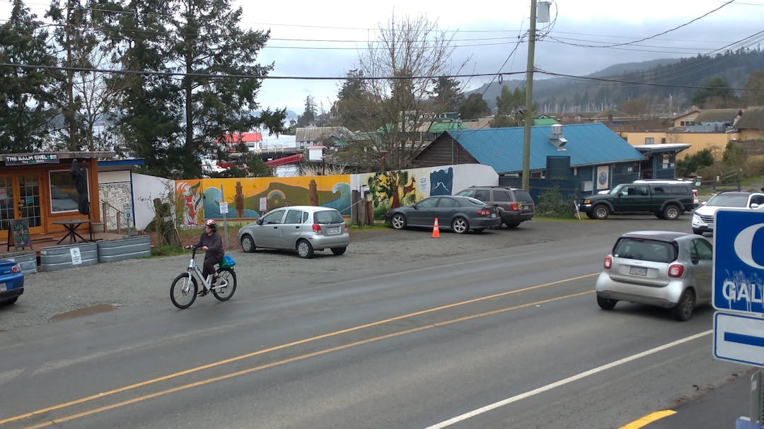 bicycle riding on road shoulder past buildings with a parked cars