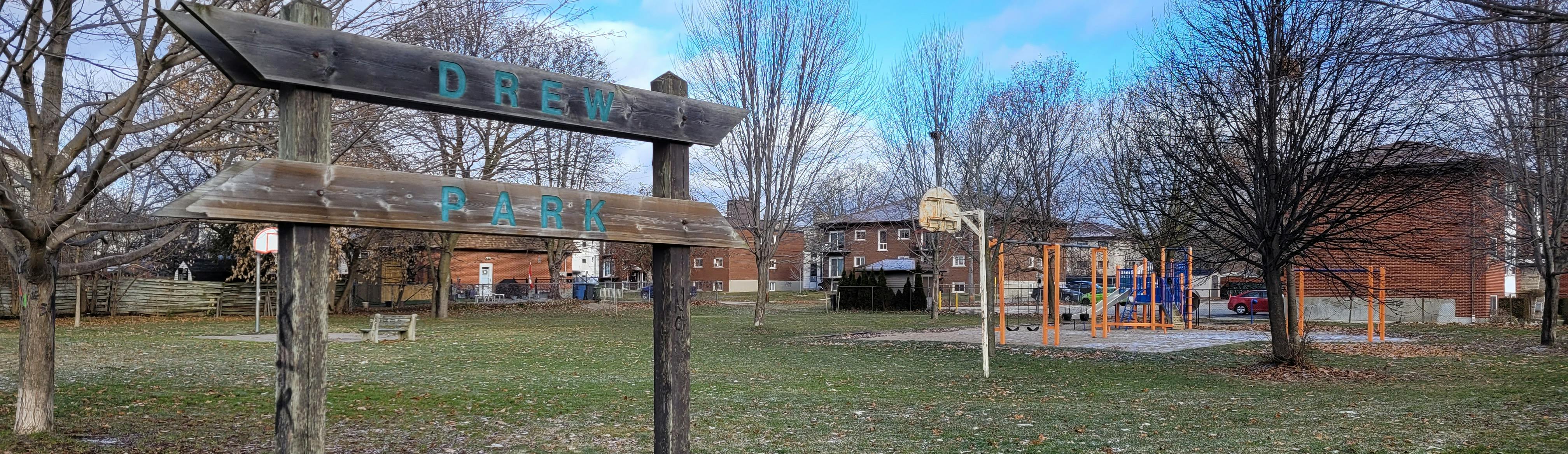 A tree lined Drew Park, showing the welcome sign, Basketball nets and playground and medium density housing in the background