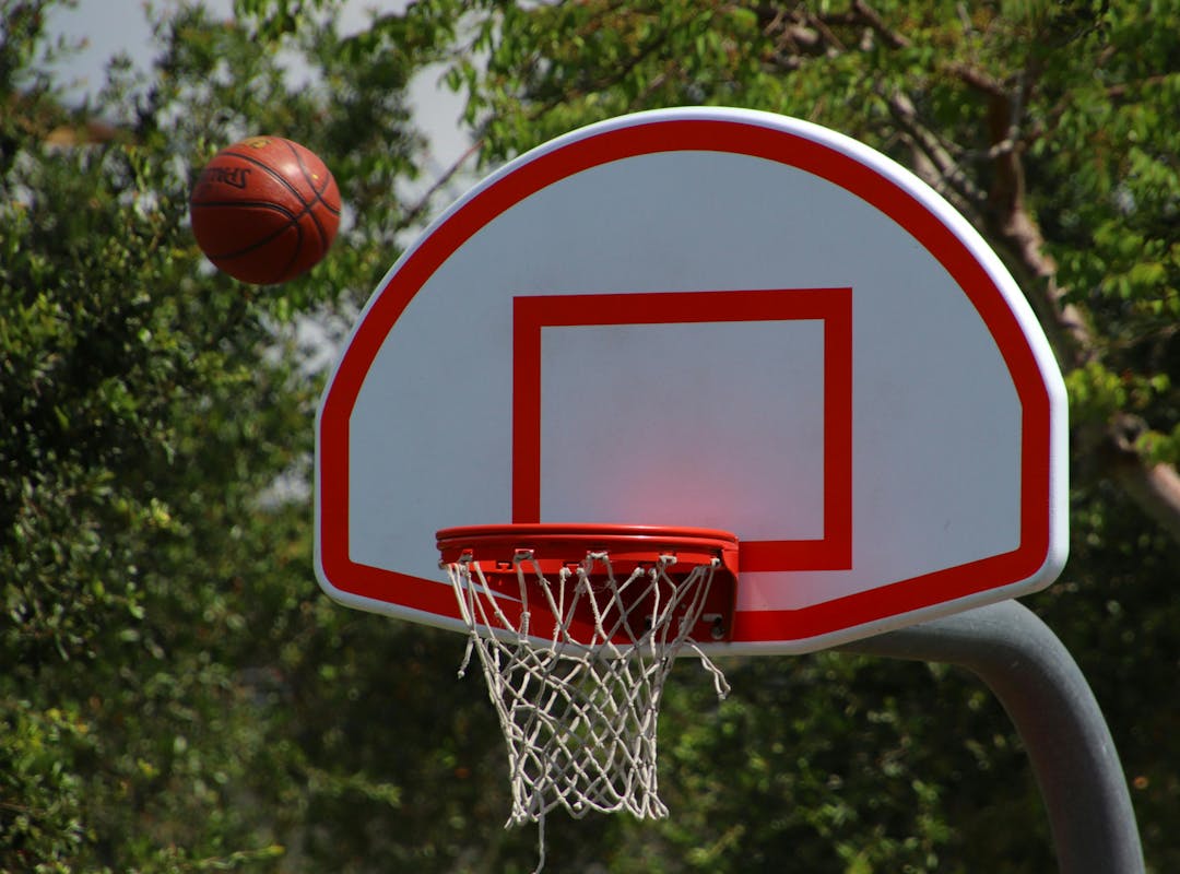 A deep orange basketball flies towards a basketball net with a white backboard and red lines, with green leafed trees behind.