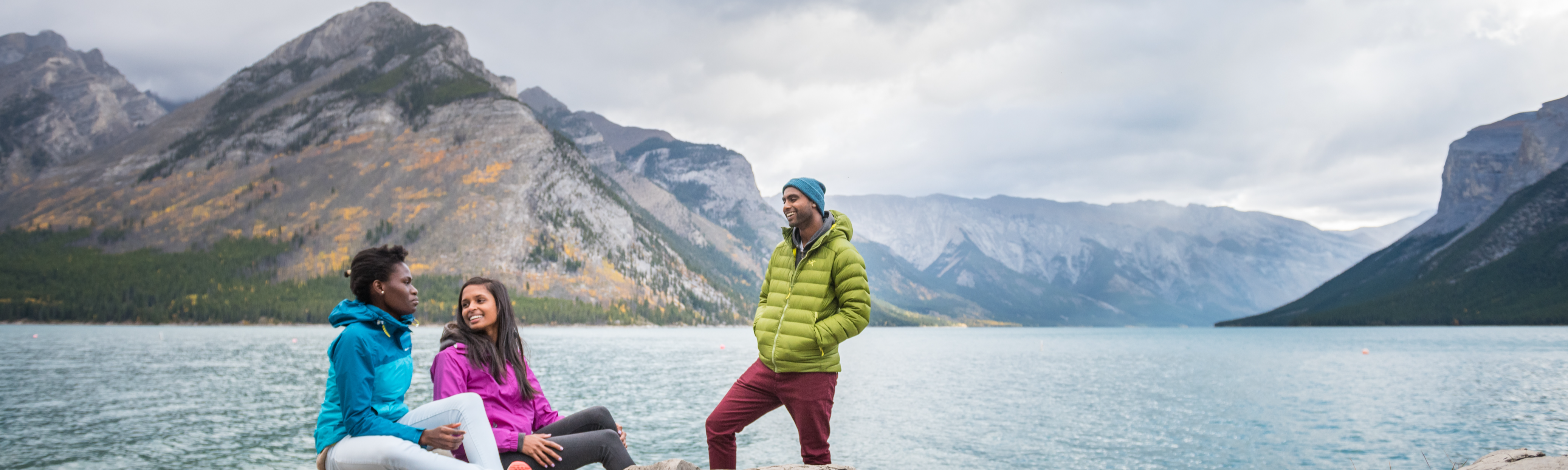 Three people chat by the shore of Lake Minnewanka on a cloudy fall day surrounded by majestic mountains.  