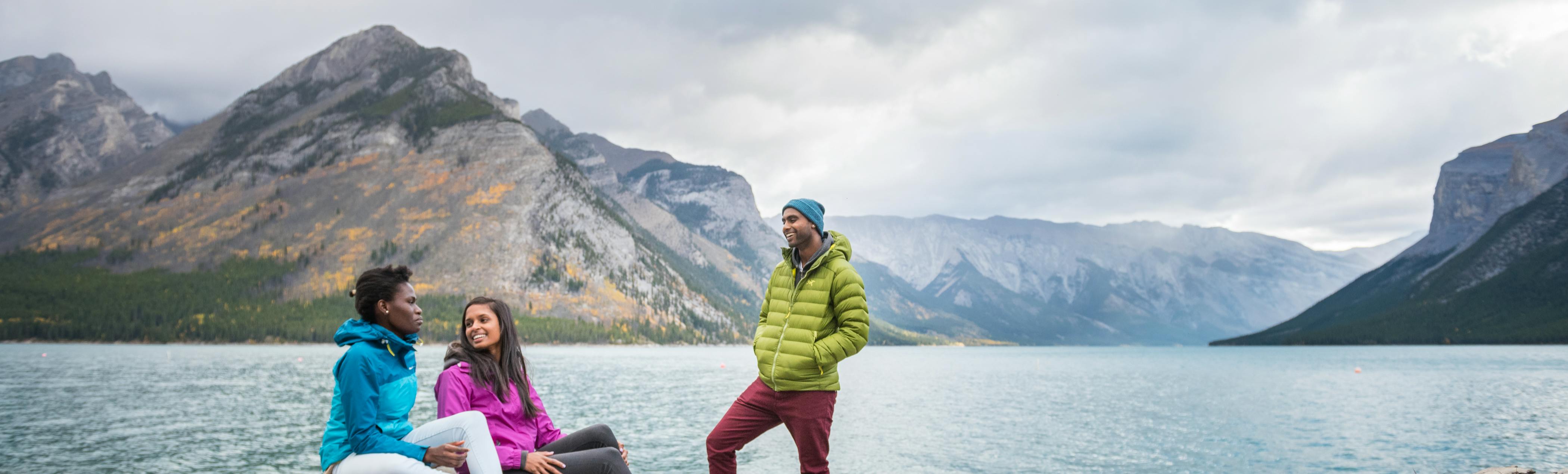 Par une journée d’automne nuageuse, trois personnes discutent au bord du lac Minnewanka, entourées de montagnes majestueuses.