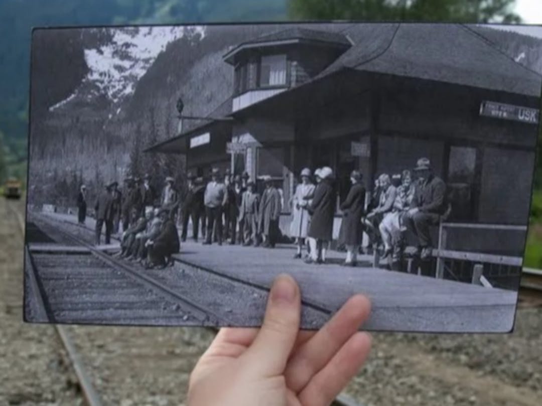 A hand holding a black and white photo of a railway station in front of the same location in present day.