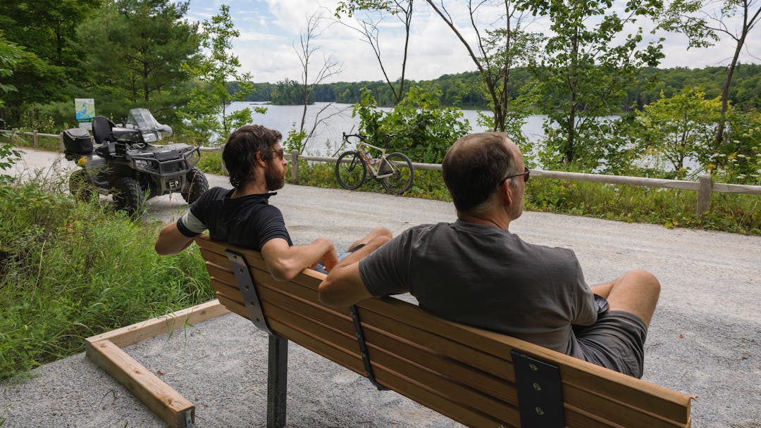 Two people sitting on a bench along a trail with an ATV and bicycle parked nearby.  A lake is in the distance.