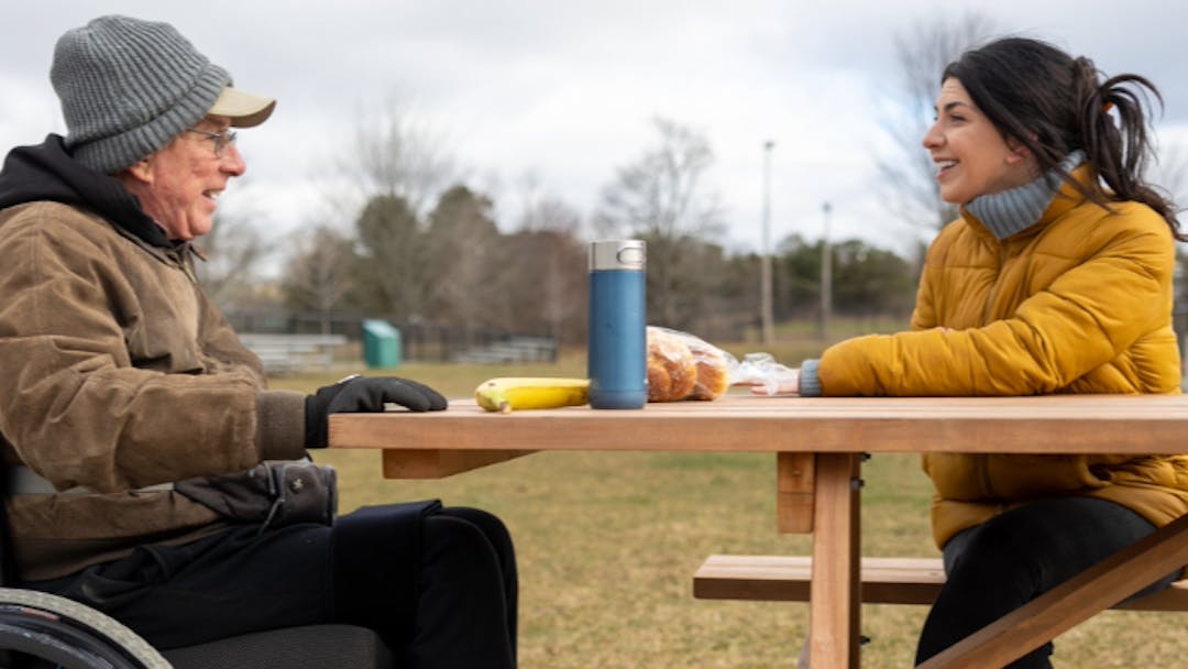 A woman is seated on a picnic table smiling and listening to an older man in a wheel chair talking with her.