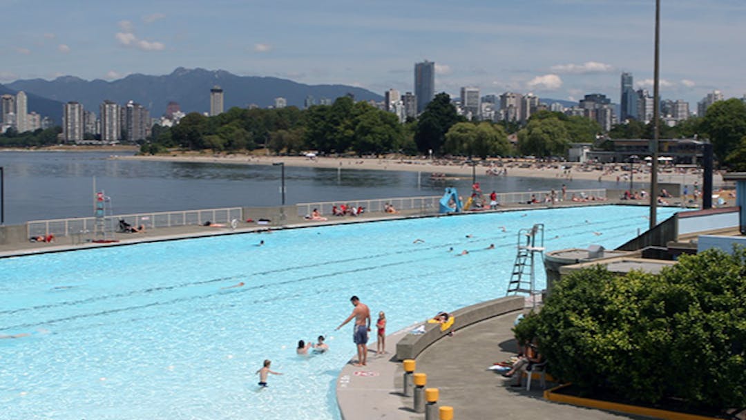 People swim at Kits Pool on a summer day, with views of the ocean and North Shore mountains in the background.  