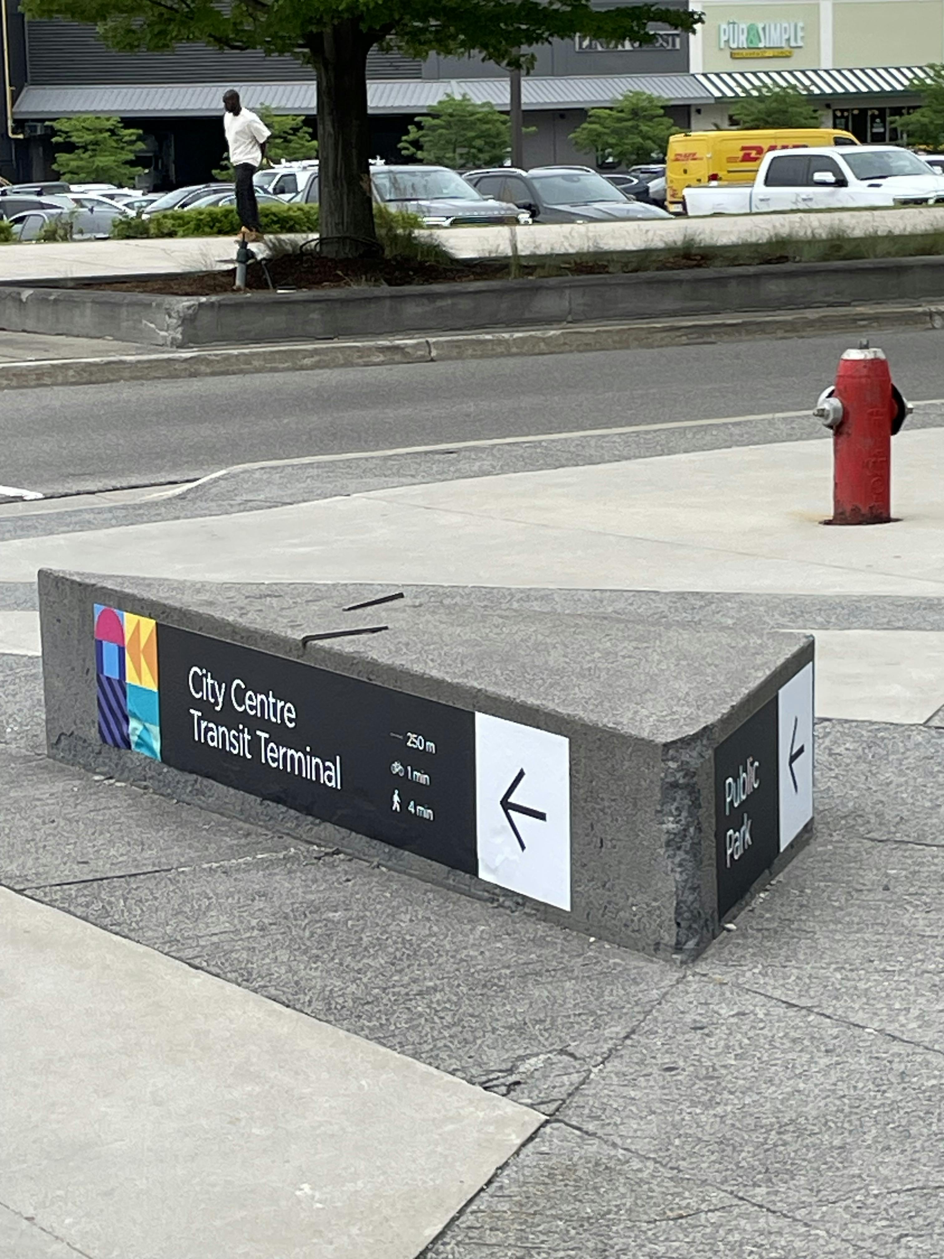 Signage on a concrete bench pointing to City Centre Transit Terminal and a public park, with a pedestrian and vehicles in the background.