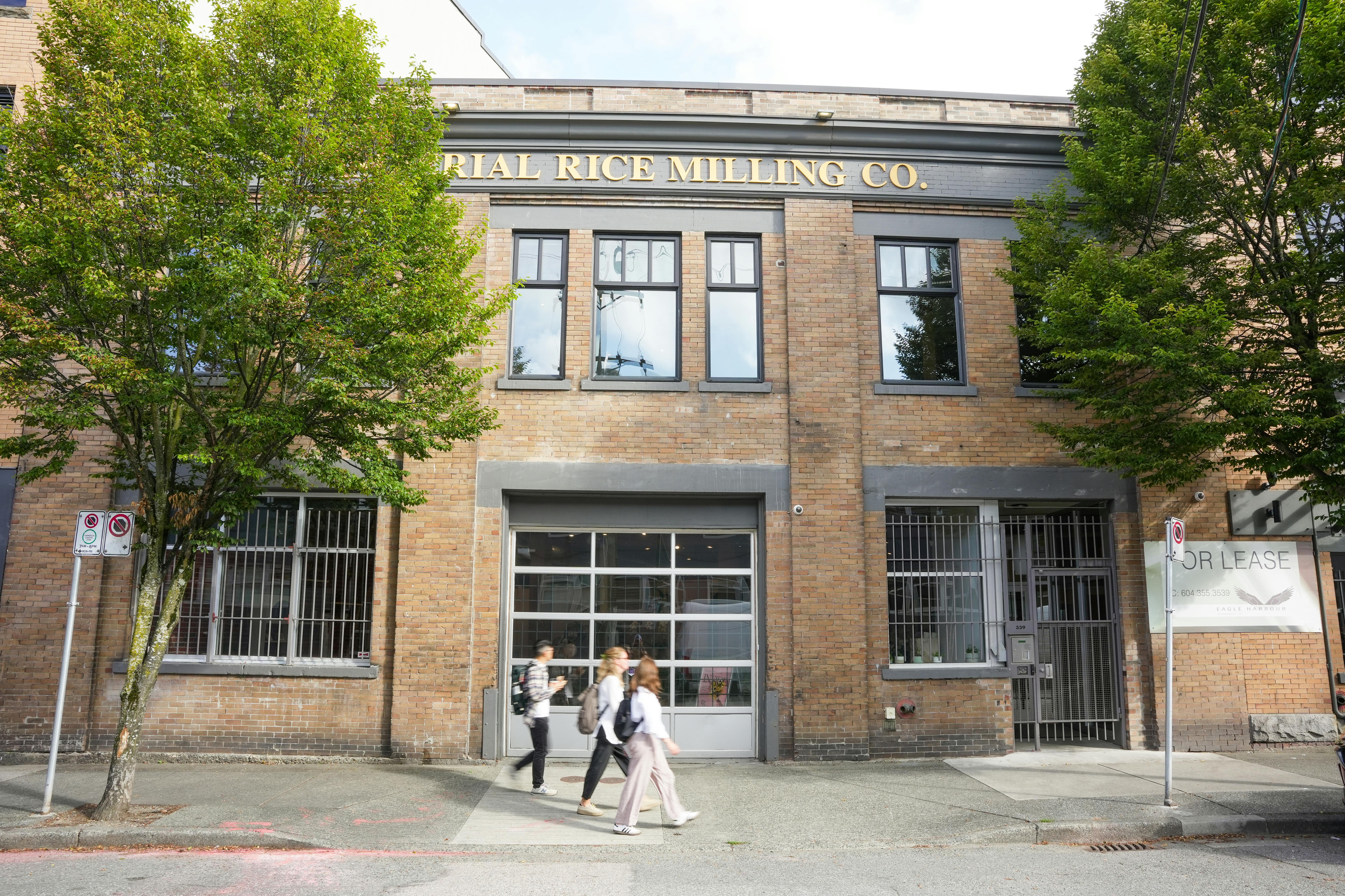Two-story brick building with large windows and a gold sign reading “Imperial Rice Milling Co.” above the second floor. Trees frame the facade, a “For Lease” sign is posted on the right, and pedestrians walk along the sidewalk.