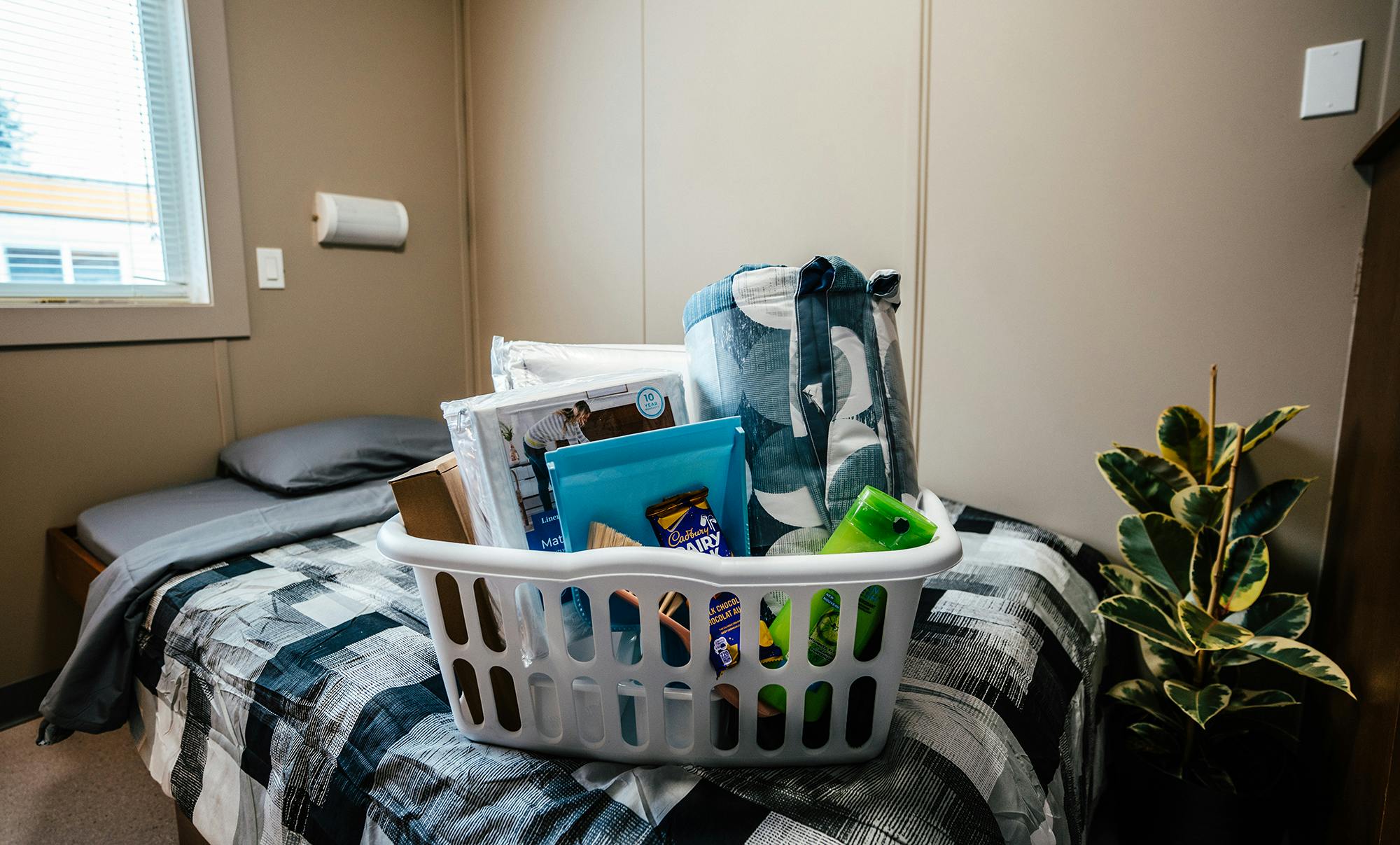 A close-up of a bed with a checkered bedspread, a window, and a laundry basket filled with personal care products, bedding, and snacks.