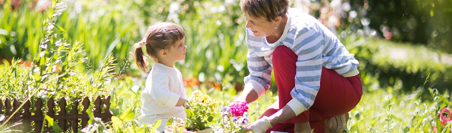 Small, female child gardening with senior in sunny garden, both looking at one another smiling.