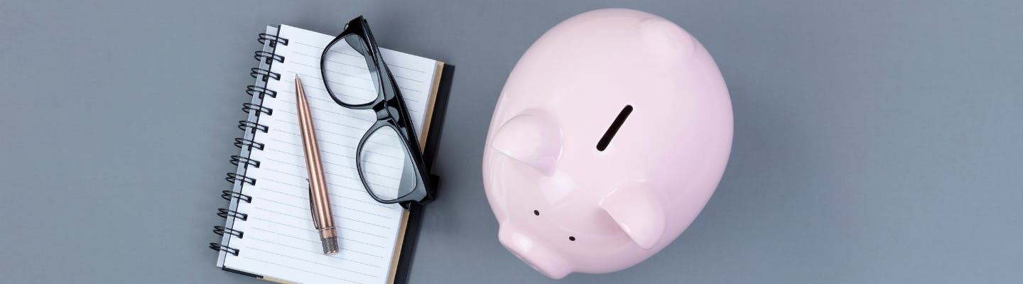 A notepad sits with glasses and pen on top. A piggy bank sits beside it. 