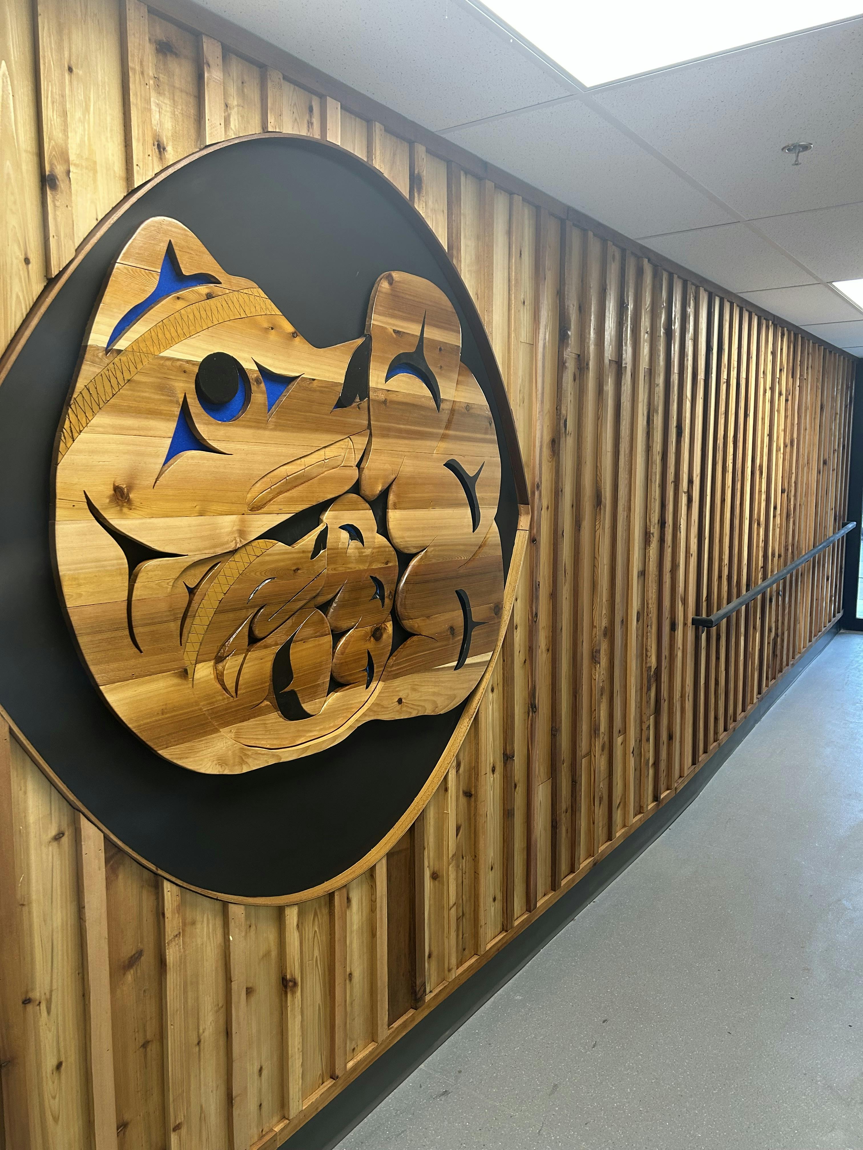 A wooden Indigenous-style artwork featuring a stylized animal design on a wood-paneled wall in a hallway.