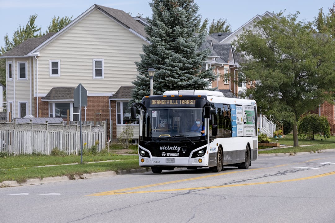 A white bus branded as Orangeville Transit stops on the side of a residential road with trees and houses in the background/