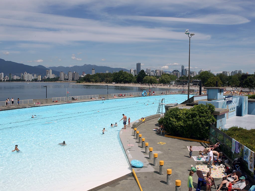 People at Kits Pool on a sunny day with some swimming and others lounging, with the mountains, city, and Showboat community stage in the background.