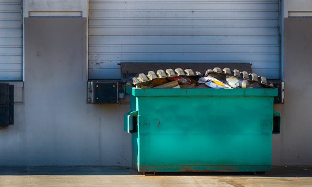 Image of the back of a commercial building with a loading dock and a large green waste dumpster sitting on the concrete.