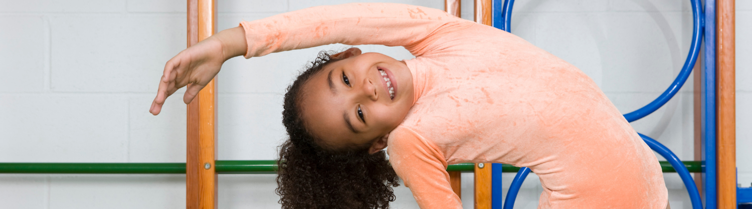 A smiling young girl practices stretches in front of gymnastics equipment
